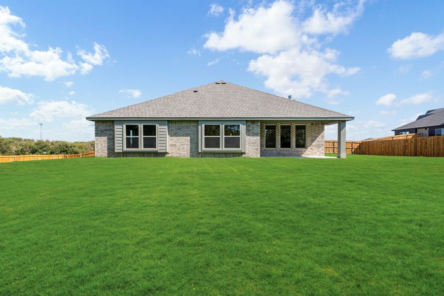 Rear view of single-story brick home with covered patio, large fenced backyard, and green lawn