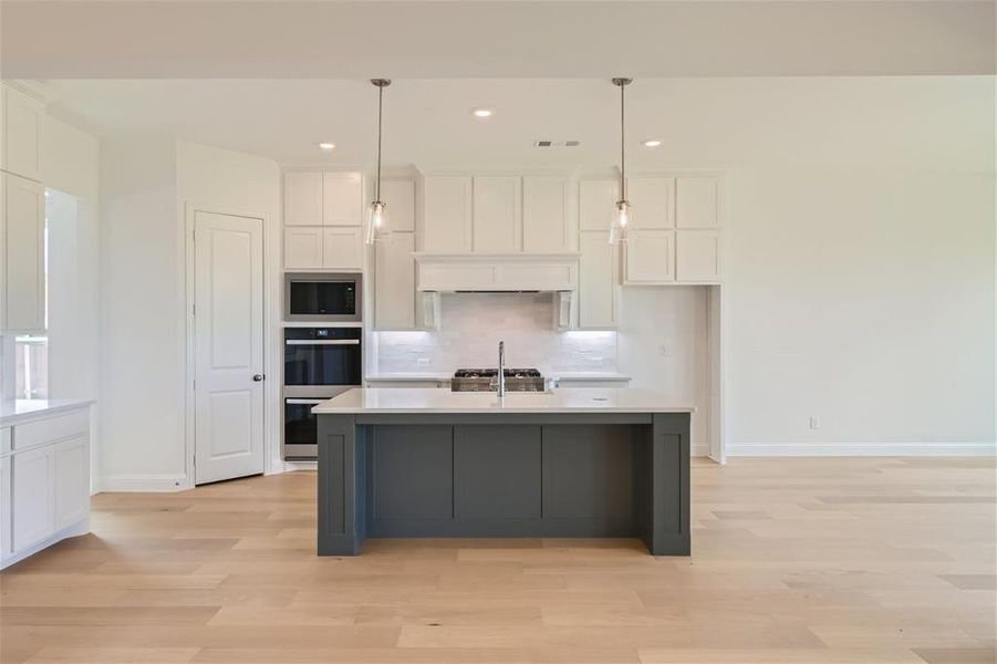 Kitchen featuring light wood-type flooring, stainless steel appliances, an island with sink, backsplash, and pendant lighting