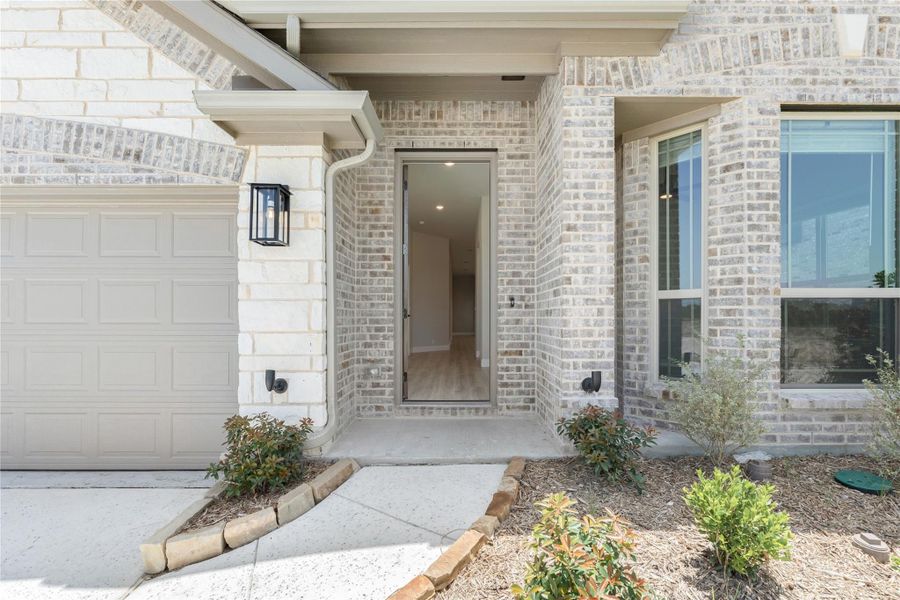Front entry of a brick home with open door, covered porch, black wall lantern, and attached garage nearby.