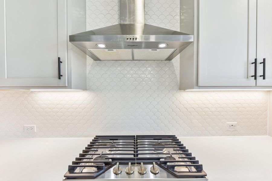 Modern kitchen features a gas range, sleek range hood, and captivating scalloped tile backsplash.