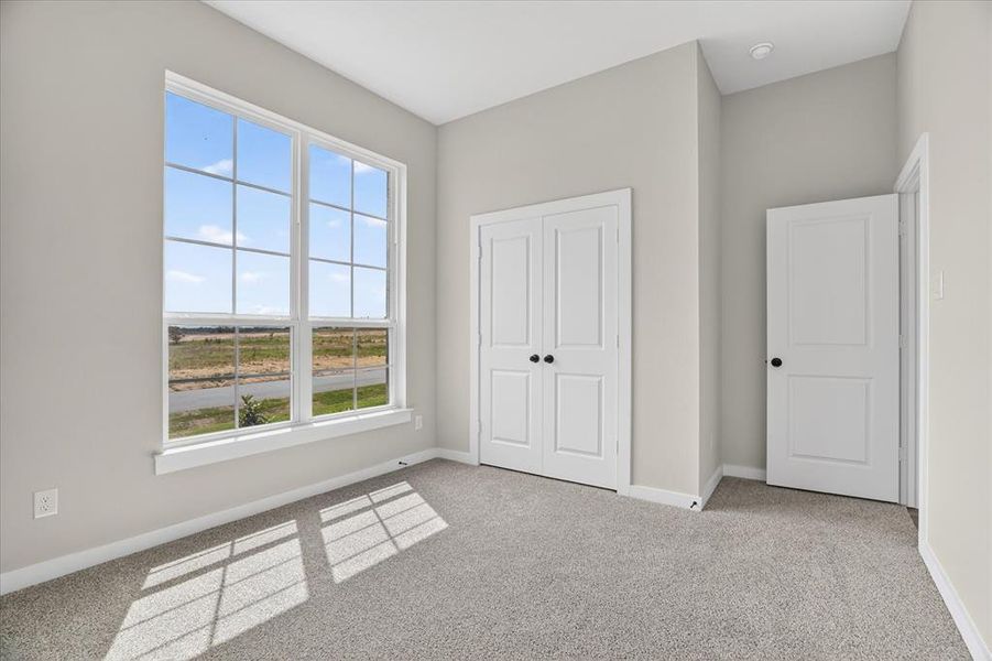 Light-filled secondary bedroom featuring a large multi-pane window, neutral-toned walls, plush grey carpeting, white trim, and two paneled doors with dark hardware