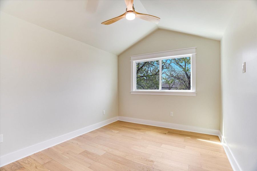 Room featuring hardwood floors, a vaulted ceiling, a ceiling fan, and a window providing views of trees Room featuring hardwood floors, a vaulted ceiling, a ceiling fan, and a window providing views of trees
