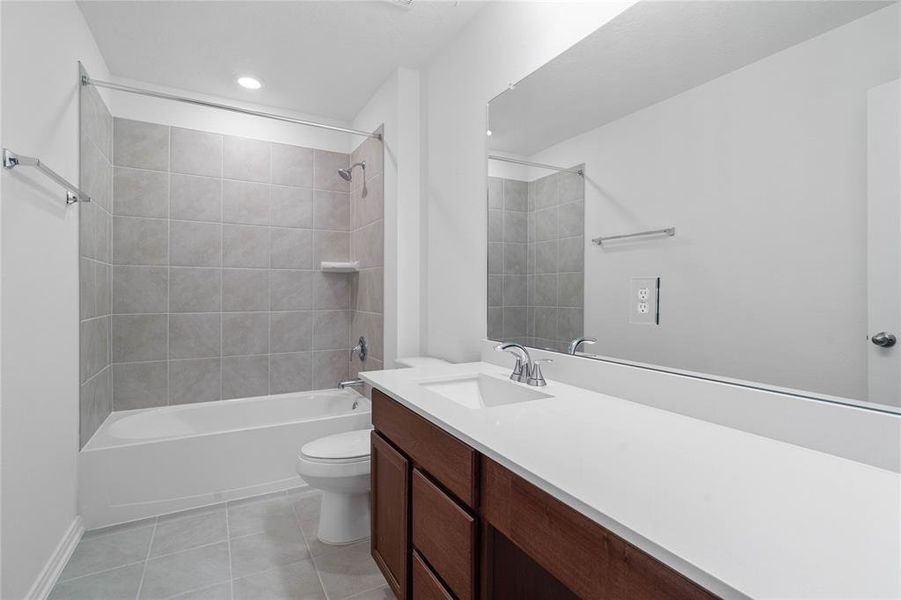 Secondary bath features tile flooring, bath/shower combo with tile surround, stained wood cabinets, beautiful light countertops, mirror, dark, sleek fixtures and modern finishes.