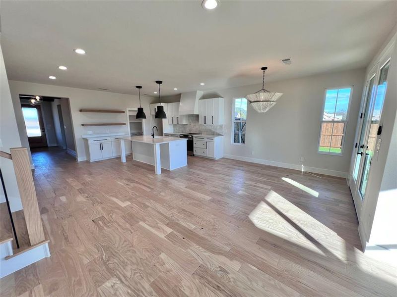 View from Living to Kitchen. Designer Light Fxture, Light Colored Hardwood Flooring, large serving  island. Floating shelves