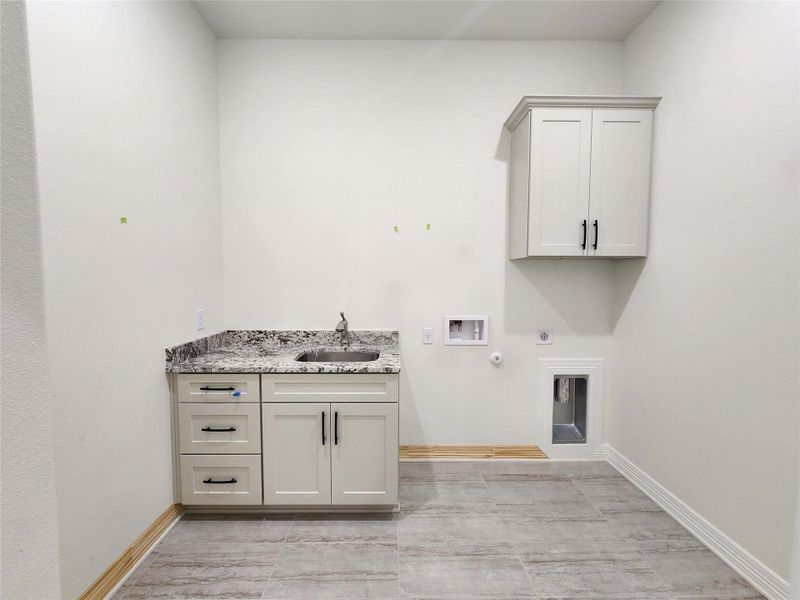 Utility room featuring light grey cabinetry with black hardware, granite countertop with integrated sink, and grey tile flooring