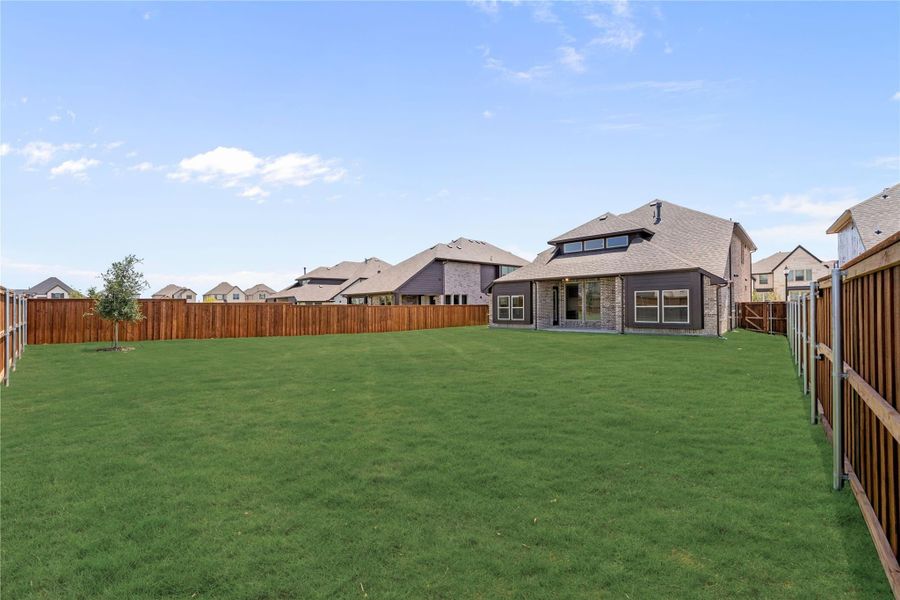 Large backyard with green lawn, wood privacy fence, and brick home with covered patio in suburban neighborhood