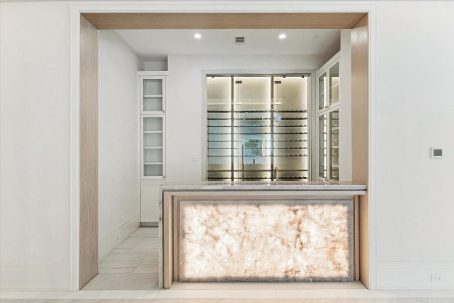 Sophisticated bar area featuring a backlit translucent stone panel, quartz countertop, and a glass-enclosed wine display with illuminated bottle racks accommodating up to 250 bottles. Flanked by built-in glass-front cabinets and framed with warm wood accents and tall cased openings.