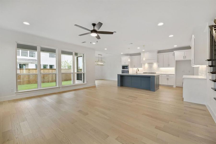 Open-concept living area featuring light wood-finish flooring, a contemporary ceiling fan with integrated lighting, and multiple large windows overlooking a private fenced yard Open-concept living area featuring light wood-finish flooring, a contemporary ceiling fan with integrated lighting, and multiple large windows overlooking a private fenced yard