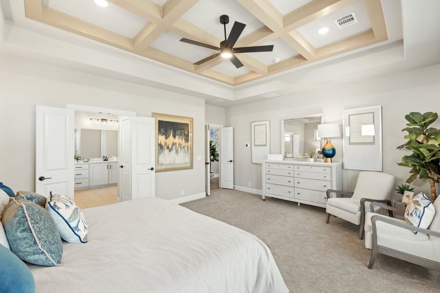 Master bedroom with coffered ceiling, ceiling fan, white dresser, and ensuite bathroom visible through open doors Master bedroom with coffered ceiling, ceiling fan, white dresser, and ensuite bathroom visible through open doors