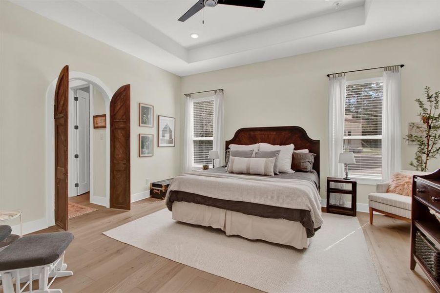 This is a spacious and well-lit primary bedroom featuring large windows, a ceiling fan, hardwood floors, and elegant double doors leading to an ensuite bathroom.