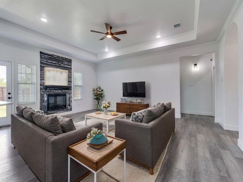 Living area featuring a raised ceiling, crown molding, wood finished floors, ceiling fan, and a fireplace Living area featuring a raised ceiling, crown molding, wood finished floors, ceiling fan, and a fireplace