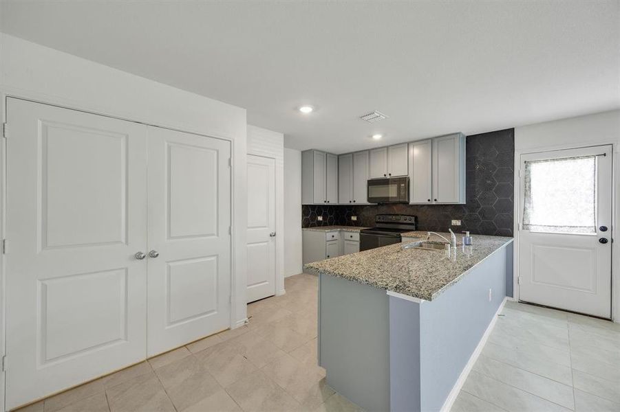 Kitchen featuring light grey cabinetry, black hexagon tile backsplash, black built-in appliances, a granite countertop island with a stainless steel sink, and light tile flooring