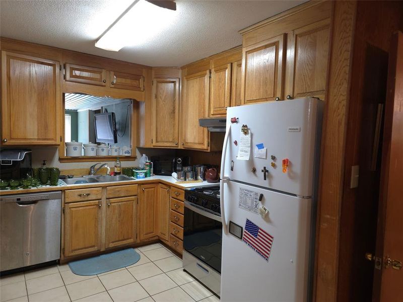 Kitchen featuring freestanding refrigerator, stainless steel dishwasher, gas range, light tile patterned floors, and brown cabinetry Kitchen featuring freestanding refrigerator, stainless steel dishwasher, gas range, light tile patterned floors, and brown cabinetry