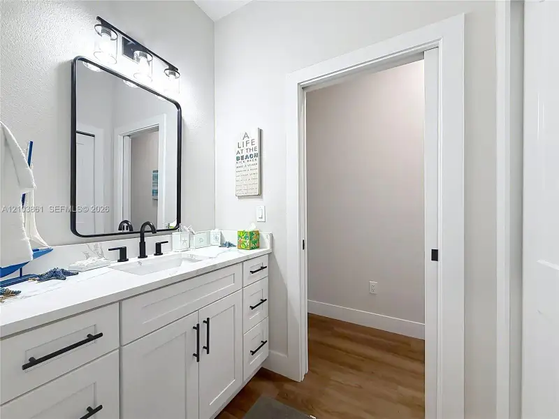 White cabinetry vanity with stone-finish countertop, undermount sink, matte black hardware, rectangular framed mirror, and wood-finish flooring