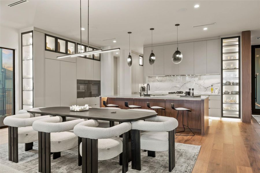 Open-concept living area featuring wood-finish flooring, a contemporary dining table, a kitchen island with seating, a built-in oven, and custom display cabinetry with integrated lighting