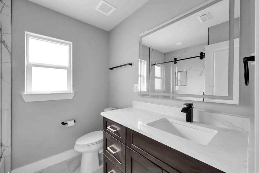 This bathroom features a modern design with a large mirror, sleek dark wood vanity, and a white countertop. It has a minimalistic look with a light color palette, a window for natural light, and a stylish black faucet and hardware.
