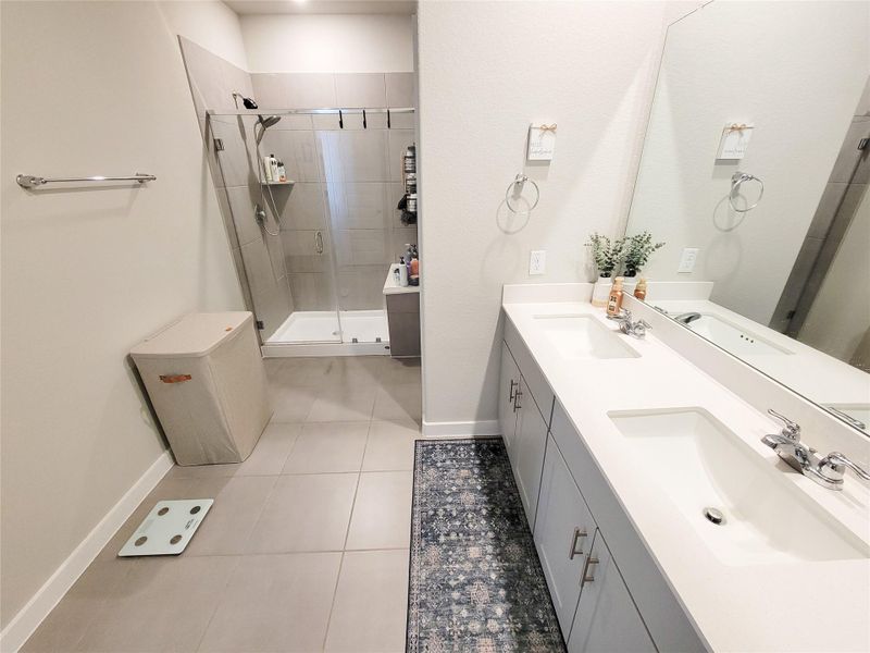 Modern bathroom featuring a double vanity with sleek white countertops, a large mirror, and ample storage. There's a spacious walk-in shower with glass doors and neutral tile flooring. The space is bright and clean, with contemporary fixtures.