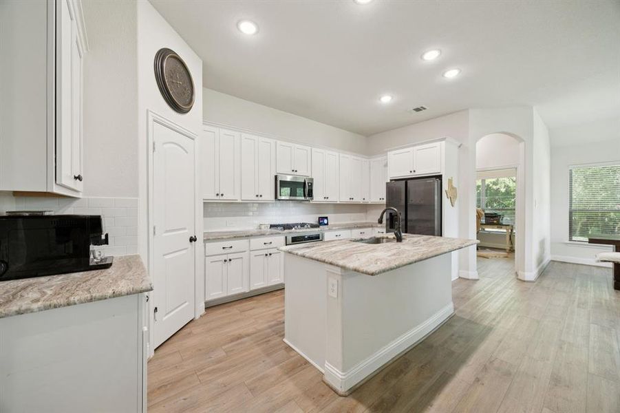 Open concept kitchen featuring white cabinets, granite counter tops, stainless steel appliances, recessed lighting, vinyl plank floors floors, and an island with sink.
