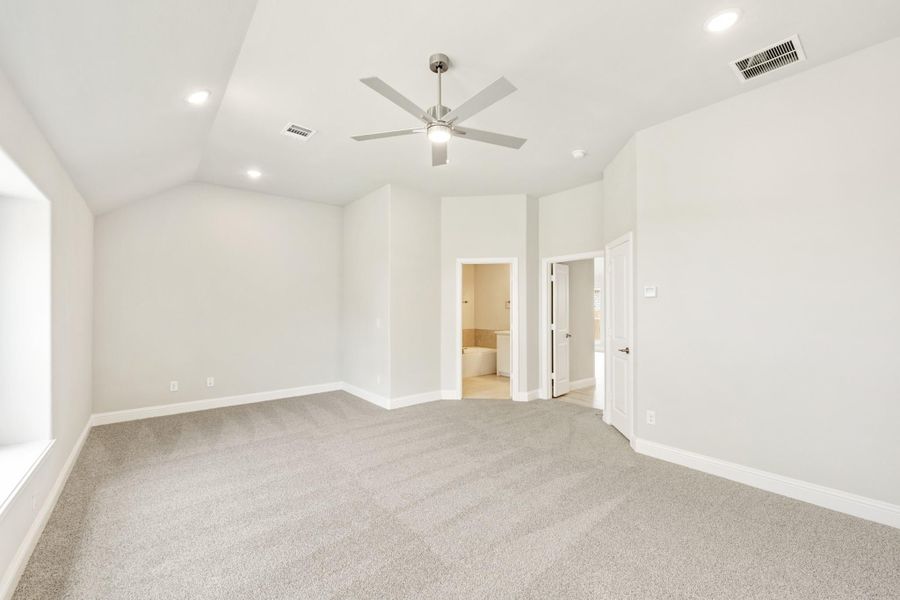 Spacious bedroom with vaulted ceiling, ceiling fan, carpet flooring, and ensuite bathroom doorway. Spacious bedroom with vaulted ceiling, ceiling fan, carpet flooring, and ensuite bathroom doorway.