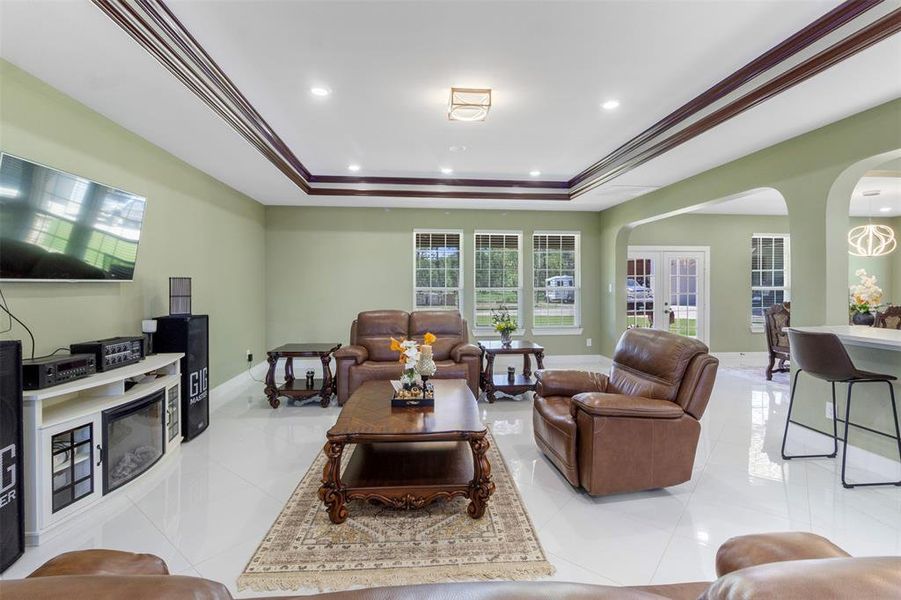 Spacious living room with elegant tray ceiling, natural light from large windows, and a cozy seating area with leather furniture. It features a modern entertainment setup and an open layout leading to a dining area, perfect for family gatherings.