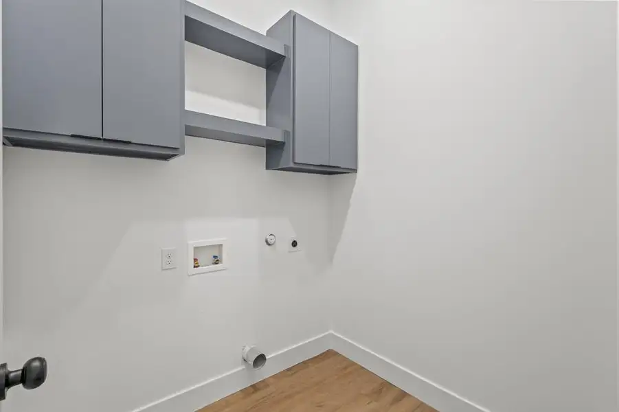 Utility space featuring overhead cabinetry in a matte gray finish, an open shelf, wood-finish flooring, and crisp white baseboards Utility space featuring overhead cabinetry in a matte gray finish, an open shelf, wood-finish flooring, and crisp white baseboards