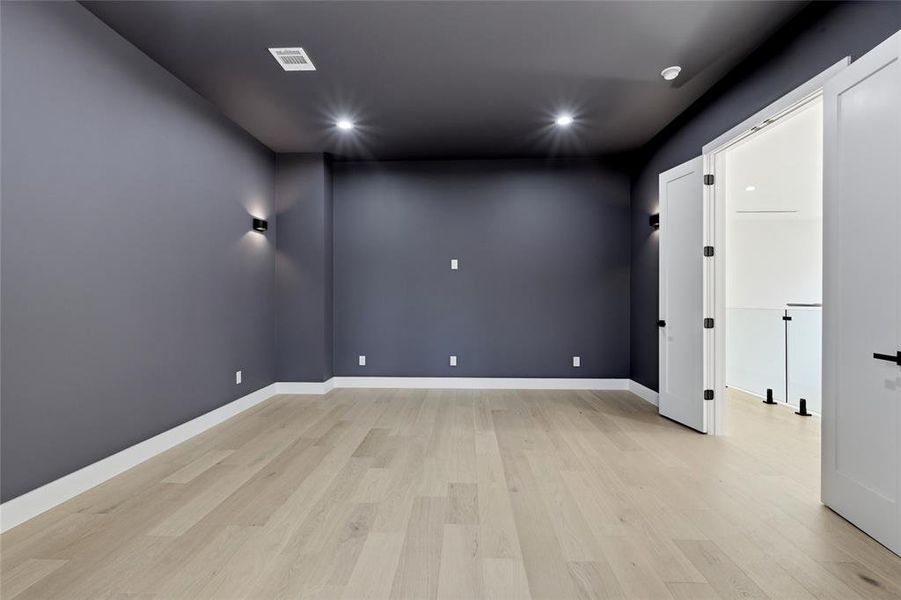Spacious room with light wood-finish flooring, dark accent walls, recessed lighting, wall sconces, and double doors opening to an adjacent area with a glass railing Spacious room with light wood-finish flooring, dark accent walls, recessed lighting, wall sconces, and double doors opening to an adjacent area with a glass railing