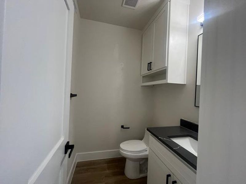 Compact bathroom featuring a white toilet, contemporary vanity with a dark countertop and integrated sink, white cabinetry with black hardware, and wood-finish flooring Compact bathroom featuring a white toilet, contemporary vanity with a dark countertop and integrated sink, white cabinetry with black hardware, and wood-finish flooring