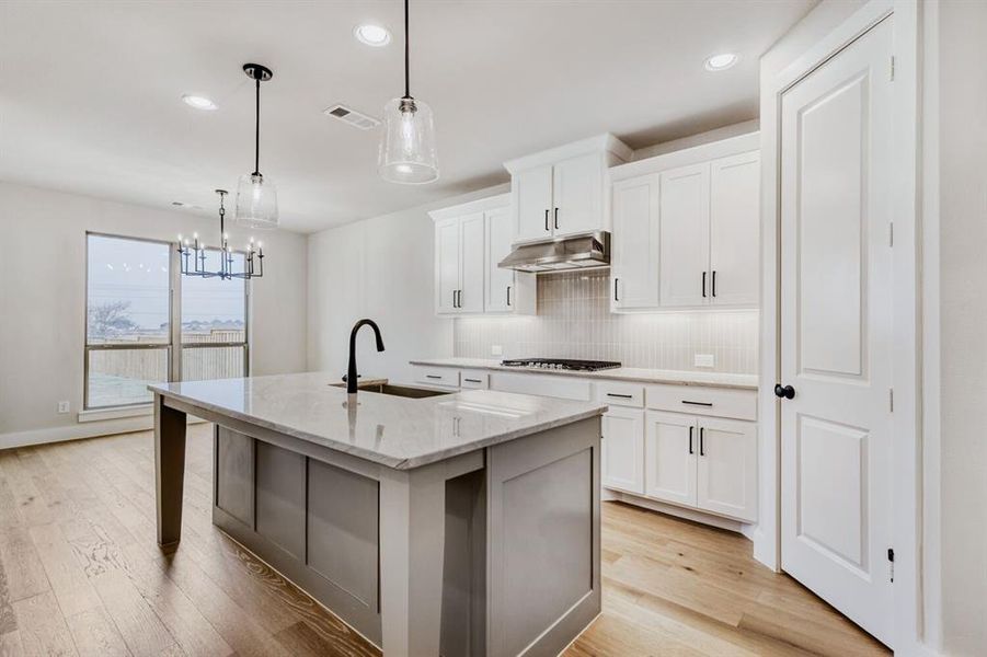 Kitchen with light wood-type flooring, decorative backsplash, an island with sink, light stone counters, and a chandelier