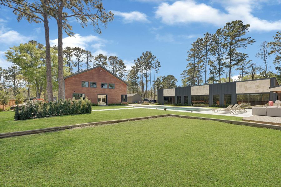 Modern home with a spacious, well-manicured lawn and a sleek, contemporary design. The property is surrounded by tall trees, offering a serene and private setting. The expansive outdoor area includes a wooden deck with seating, ideal for relaxation or entertaining.