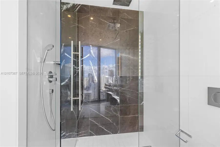 Walk-in shower featuring large-format dark stone tile accent wall, glass enclosure, ceiling-mounted rainfall showerhead, and integrated chrome handheld sprayer