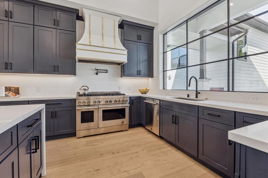 This kitchen features dark cabinetry, a large stainless steel range, a custom hood, sleek countertops, and a spacious window that provides ample natural light.