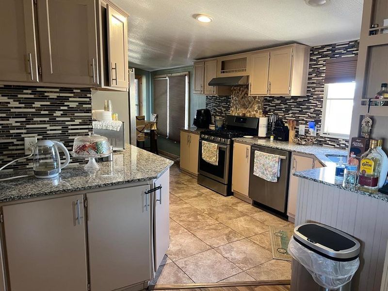 Kitchen featuring backsplash, ventilation hood, light stone countertops, light tile patterned floors, and stainless steel appliances