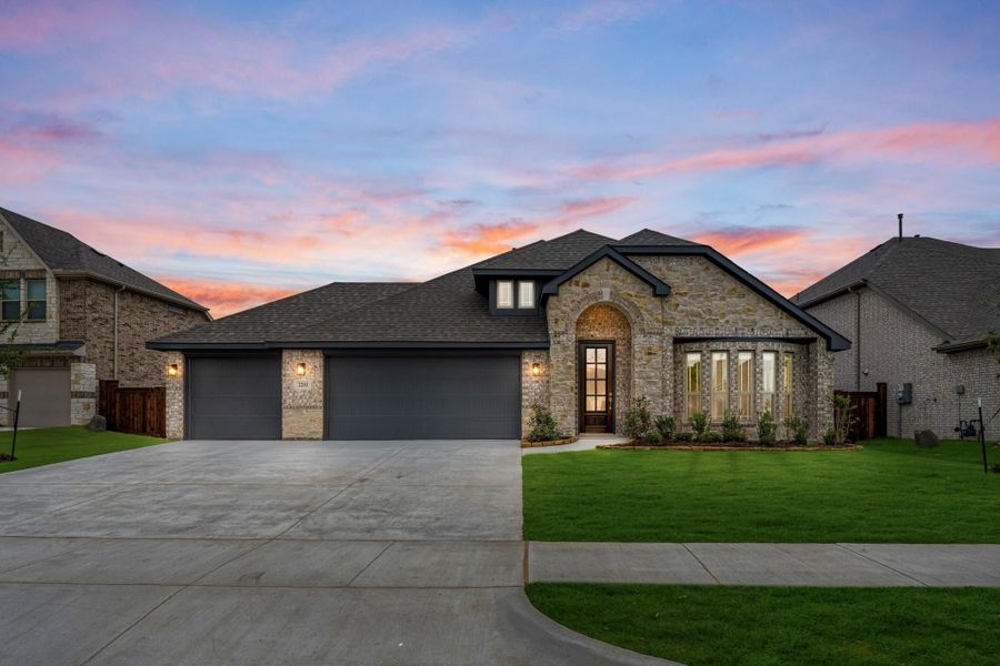 Single-story stone and brick home with three-car garage, arched entry, and green front lawn at dusk. Single-story stone and brick home with three-car garage, arched entry, and green front lawn at dusk.