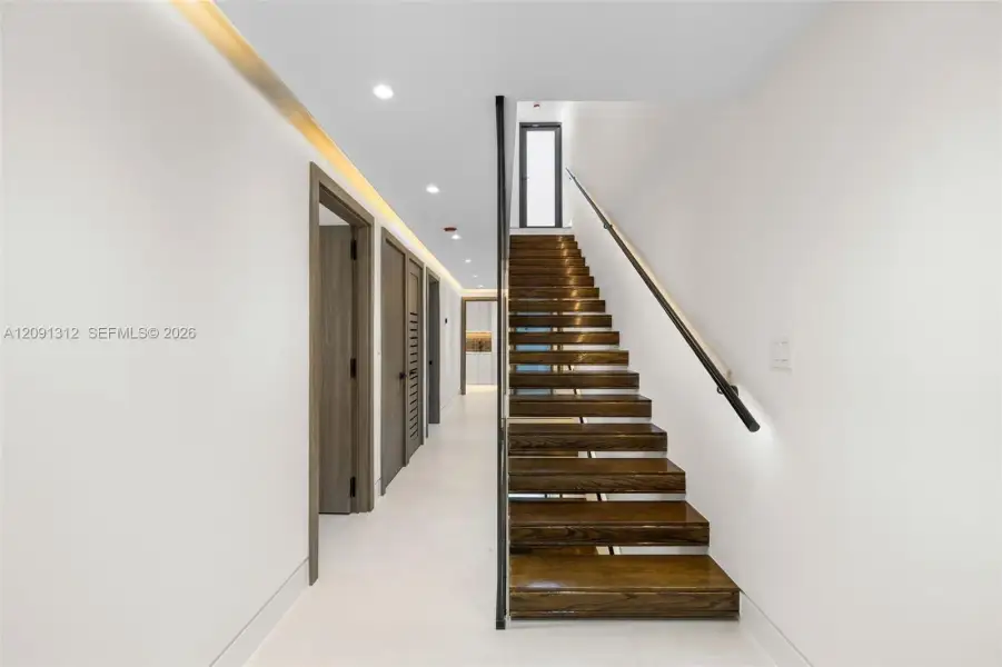 Open-tread staircase featuring wood-finish steps, a minimalist black metal handrail, recessed ceiling lighting, and smooth-finish flooring Open-tread staircase featuring wood-finish steps, a minimalist black metal handrail, recessed ceiling lighting, and smooth-finish flooring