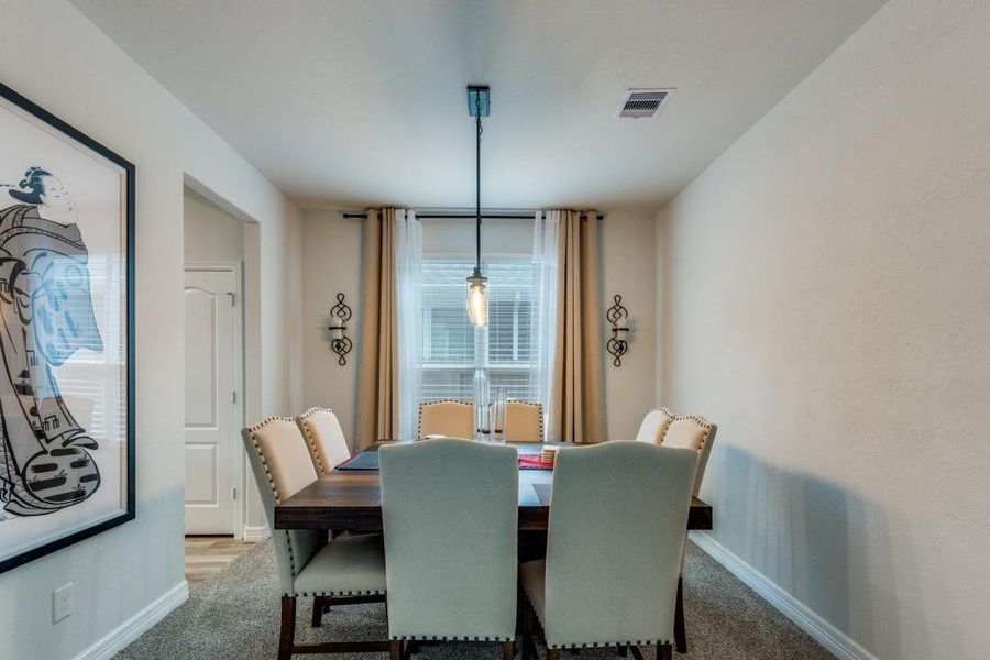 This dining room features a cozy, neutral-toned space with a modern light fixture, a large window with curtains, and a stylish wall art piece. The room includes a table with upholstered chairs, offering a comfortable setting for gatherings.