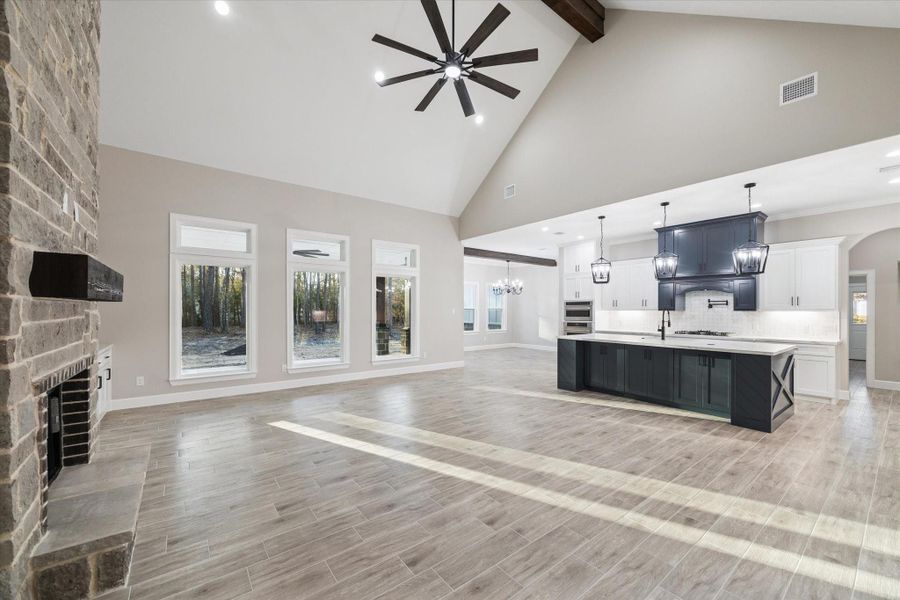 View from secondary bedroom hallway toward a wall of windows in the family room. Windows look to the rear of the home.  High beam ceiling with large lighted ceiling fan.   Builder signature design 20ft. floor to ceiling brick fireplace with dark mantle. Island kitchen with lower cabinets in designer blue.  Wood look ceramic tile flooring.