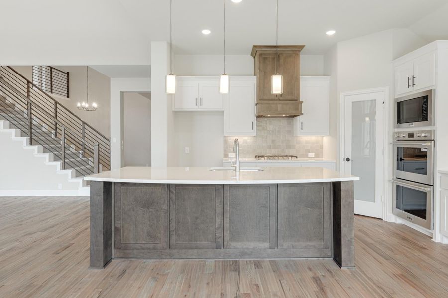 Kitchen with large gray island, white cabinets, wood range hood, double ovens, and hardwood floors Kitchen with large gray island, white cabinets, wood range hood, double ovens, and hardwood floors