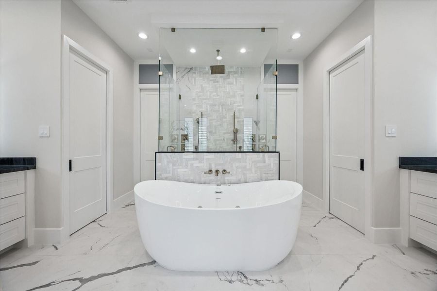 This spa-like primary bathroom is designed to impress with its modern elegance and luxurious finishes. At the center, a freestanding soaking tub creates a striking focal point, perfectly framed by a glass-enclosed shower featuring a rainfall showerhead, two hand helds and dramatic tile detail.