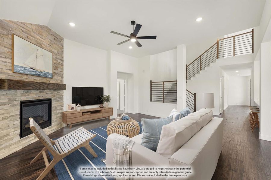 Open living room with stone fireplace, high ceilings, ceiling fan, hardwood floors, and staircase visible in background.