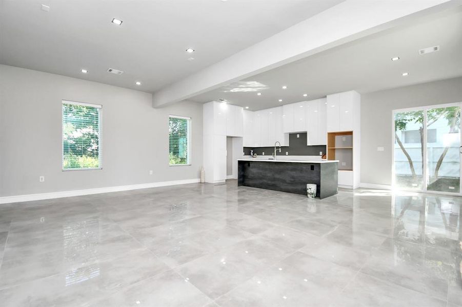 This is a spacious, modern room featuring polished tile flooring, high ceilings with recessed lighting, large windows for natural light, and a neutral color palette. The room is a blank canvas ready for personalization.