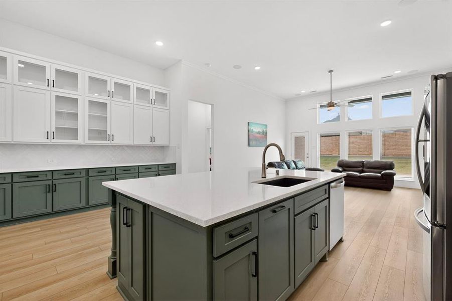 Kitchen featuring a large central island with a built-in sink and white countertop, complemented by dark green base cabinetry and white upper cabinetry with glass inserts Kitchen featuring a large central island with a built-in sink and white countertop, complemented by dark green base cabinetry and white upper cabinetry with glass inserts