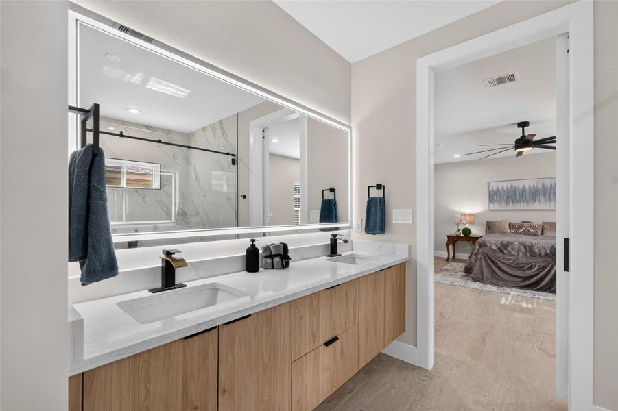 Experience modern elegance in this bathroom featuring a sleek double vanity with under-cabinet storage, a large backlit mirror, and an adjoining view into the cozy bedroom.