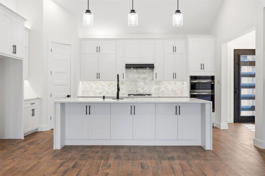 Kitchen featuring a central island with a sink, white cabinetry with dark hardware, a patterned tile backsplash, and wood-finish flooring Kitchen featuring a central island with a sink, white cabinetry with dark hardware, a patterned tile backsplash, and wood-finish flooring