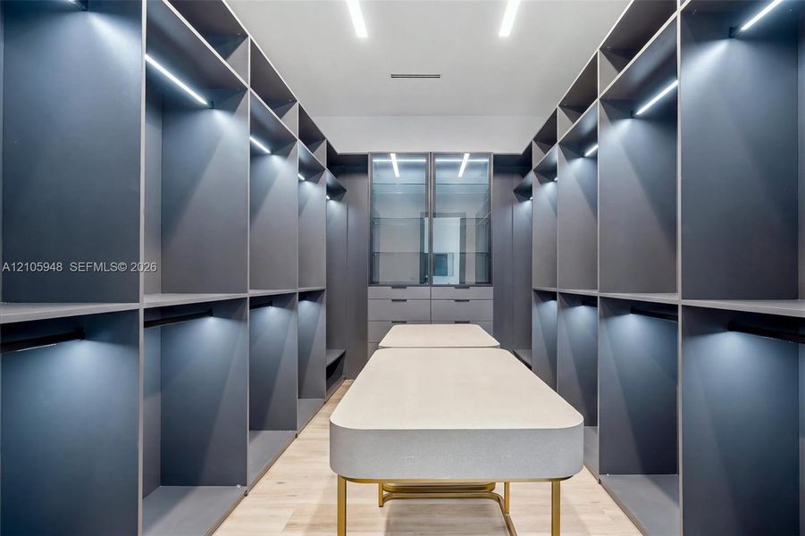 Custom walk-in wardrobe featuring floor-to-ceiling matte cabinetry, integrated LED shelving, glass-fronted display cases, a central island with a stone-finish countertop, and light-tone wood-finish flooring