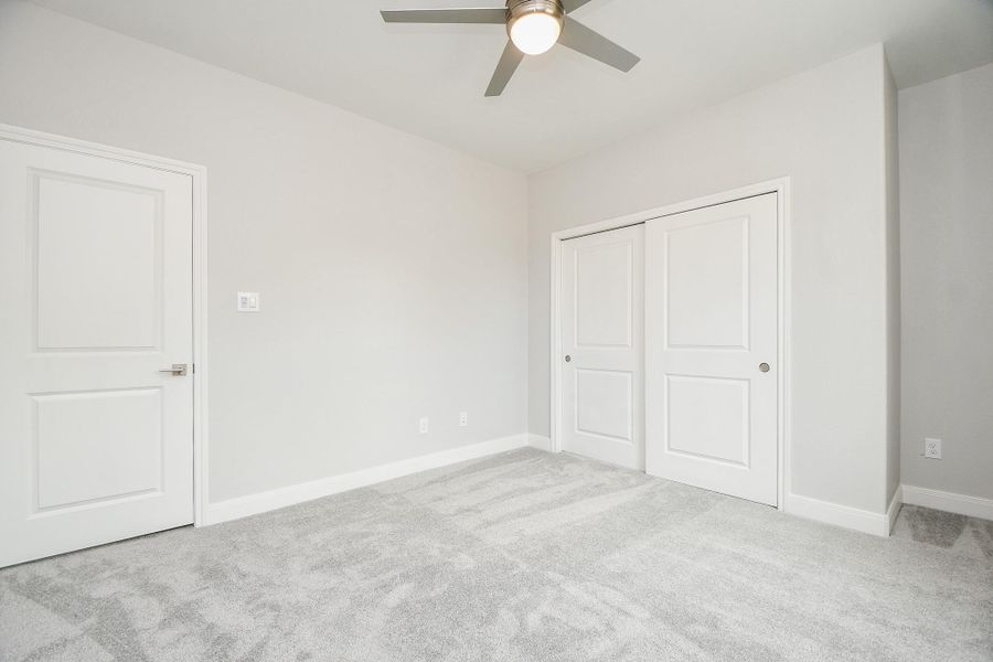 This is a bright, modern bedroom with light gray walls and carpet. It features a ceiling fan and a double-door closet, offering a clean and spacious feel.