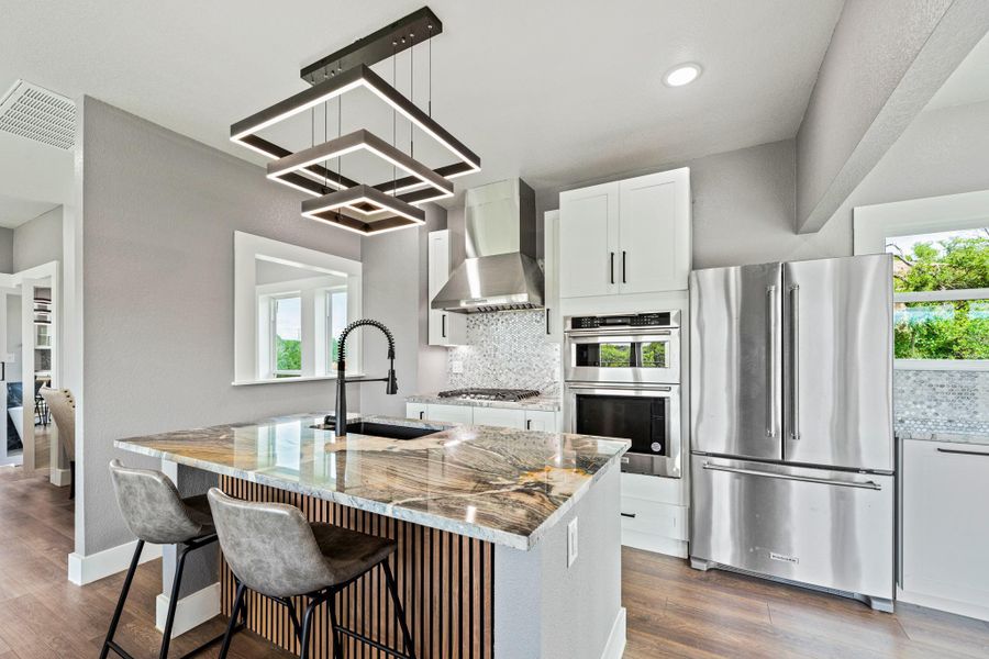 Modern kitchen featuring a striking geometric chandelier, stainless steel appliances, a large center island with a stone countertop, a tile backsplash, and white cabinetry