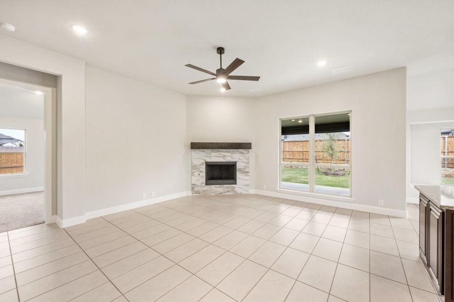 Open living room with tile floors, stone fireplace, ceiling fan, and large backyard-facing window Open living room with tile floors, stone fireplace, ceiling fan, and large backyard-facing window