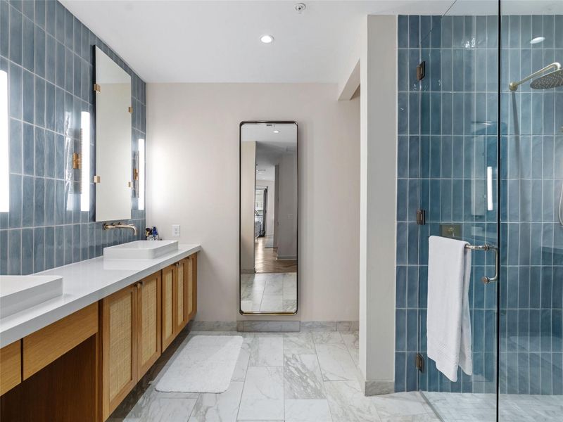 Bathroom featuring a dual vanity with rectangular vessel sinks, integrated wall-mounted mirrors with vertical lighting, light wood cabinetry with cane inserts, a full-height framed mirror, and a glass-enclosed shower with blue vertical tile and a rain showerhead Bathroom featuring a dual vanity with rectangular vessel sinks, integrated wall-mounted mirrors with vertical lighting, light wood cabinetry with cane inserts, a full-height framed mirror, and a glass-enclosed shower with blue vertical tile and a rain showerhead