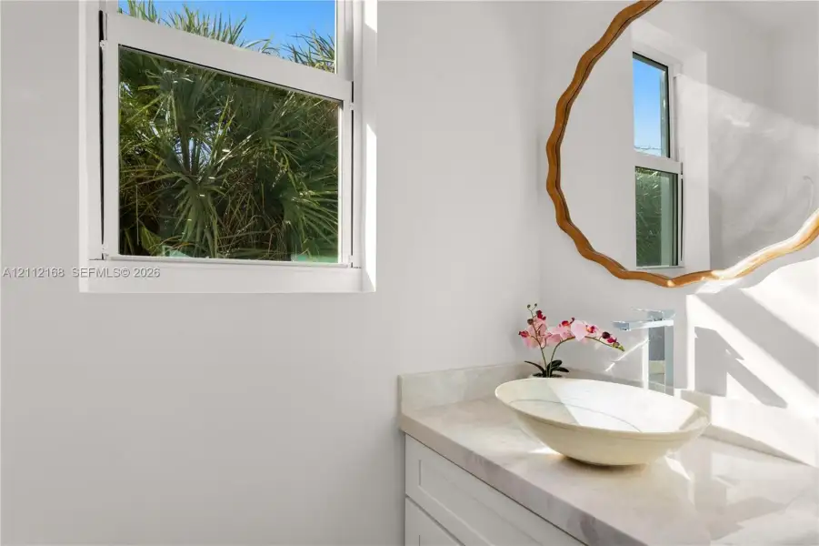 Vanity featuring a stone-surface countertop, an oval vessel sink, a chrome single-handle faucet, and white shaker-style cabinetry, complemented by a single-hung window Vanity featuring a stone-surface countertop, an oval vessel sink, a chrome single-handle faucet, and white shaker-style cabinetry, complemented by a single-hung window