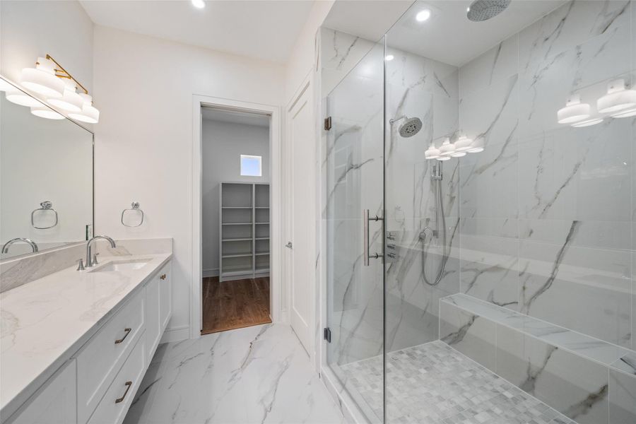 Primary bathroom features a spacious glass-enclosed shower with elegant marble-style tiles and a built-in bench. It also includes a dual-sink vanity with sleek finishes, ample lighting, and an adjacent walk-in closet with shelving.